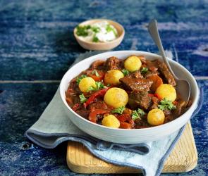 Eine Suppenschüssel mit Gulasch und Polentaklösschen und einem Löffel auf einem Holzbrett.