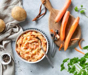 Karotten-Kartoffel-Stampf in einer Schale mit Löffel, daneben Möhren auf einem Holzbrett.
