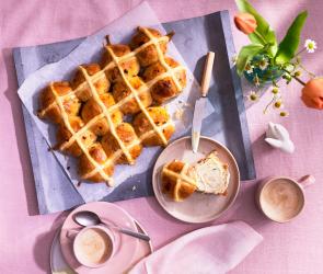 Hot Cross Buns auf einem lilafarbenen Untergrund, eins aufgeschnitten auf einem Teller.