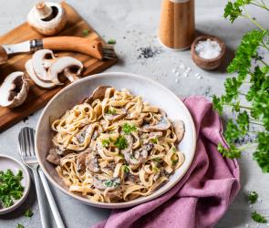 Bandnudeln mit Pilzsauce und Kräutern in einem tiefen Teller, daneben ein Brett mit Pilzen.