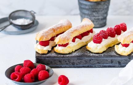 Eclairs auf Servierbrett mit Puderzucker im Sieb und Himbeeren in Schale