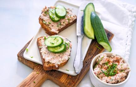 Zwei Scheiben Brot mit Streichwurst und Gurke auf Holzbrett.