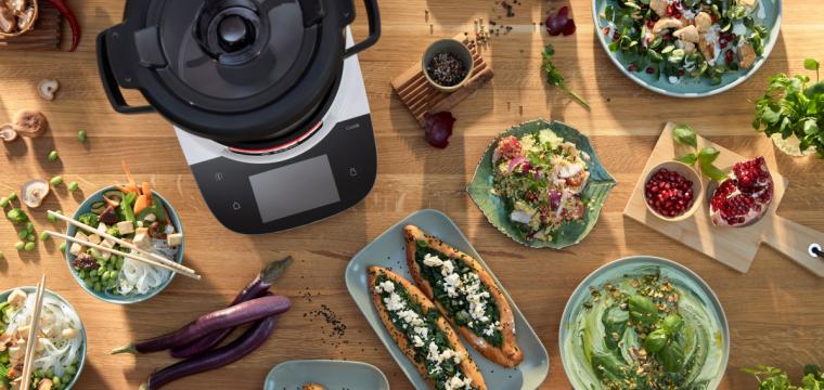 Der Cookit auf einer Tischplatte, daneben verschieden Gerichte, wie Spinat-Pita, Hummus und Couscous-Salat. 