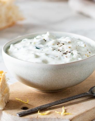 Tzatziki in Schale auf Brett mit Fladenbrot, Gurkenscheiben und weißem Tuch