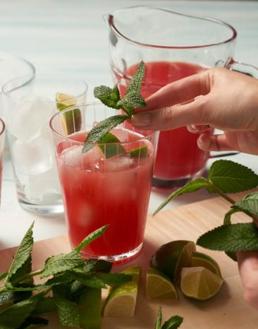 Wassermelonensaft in Gläsern und Karaffe, Limetten und Minze, Hand im Bild die Drinks dekoriert