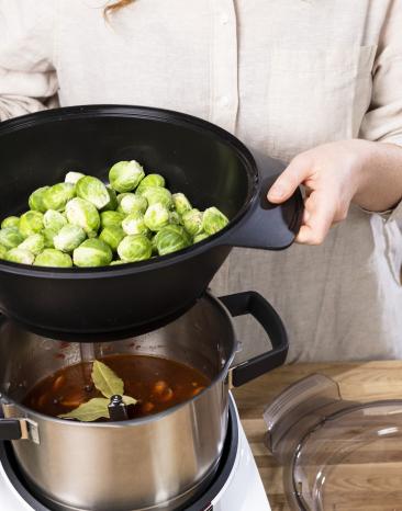 Frau setzt Dampfgaraufsatz gefüllt mit Rosenkohl auf den Cookit Kochtopf Frau setzt Dampfgaraufsatz gefüllt mit Rosenkohl auf den Cookit Kochtopf