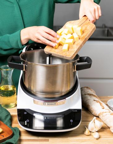 Frau schiebt Käsewürfel von einem Holzbrett in den Cookit Kochtopf Frau schiebt Käsewürfel von einem Holzbrett in den Cookit Kochtopf