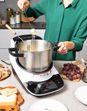 Frau zieht mit einer Fonduegabel ein mit Käse ummanteltes Stück Brot aus dem Cookit Frau zieht mit einer Fonduegabel ein mit Käse ummanteltes Stück Brot aus dem Cookit