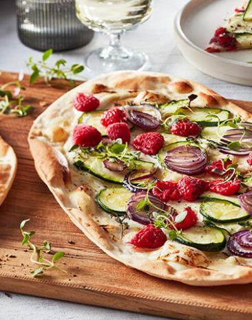 Flammkuchen belegt mit Himbeeren, Zucchini und Ziegenkäse auf einem Holzbrett.