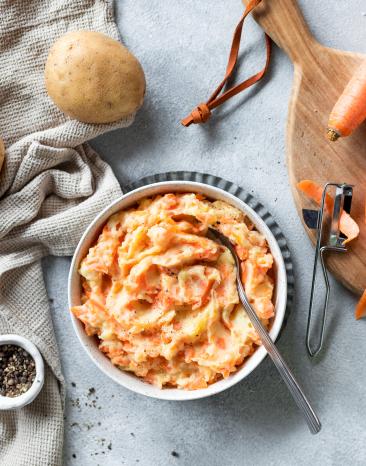 Karotten-Kartoffel-Stampf in einer Schale mit Löffel, daneben Möhren auf einem Holzbrett.