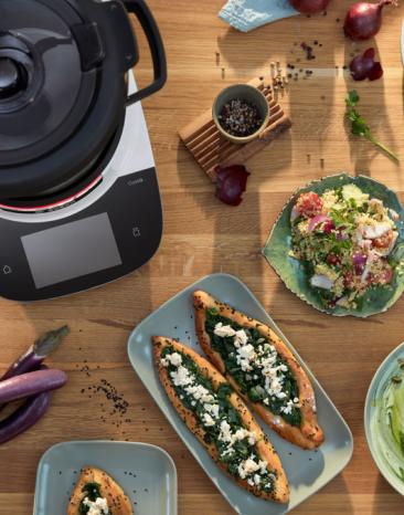 Der Cookit auf einer Tischplatte, daneben verschieden Gerichte, wie Spinat-Pita, Hummus und Couscous-Salat. 