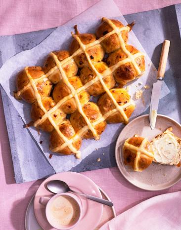 Hot Cross Buns auf einem lilafarbenen Untergrund, eins aufgeschnitten auf einem Teller.